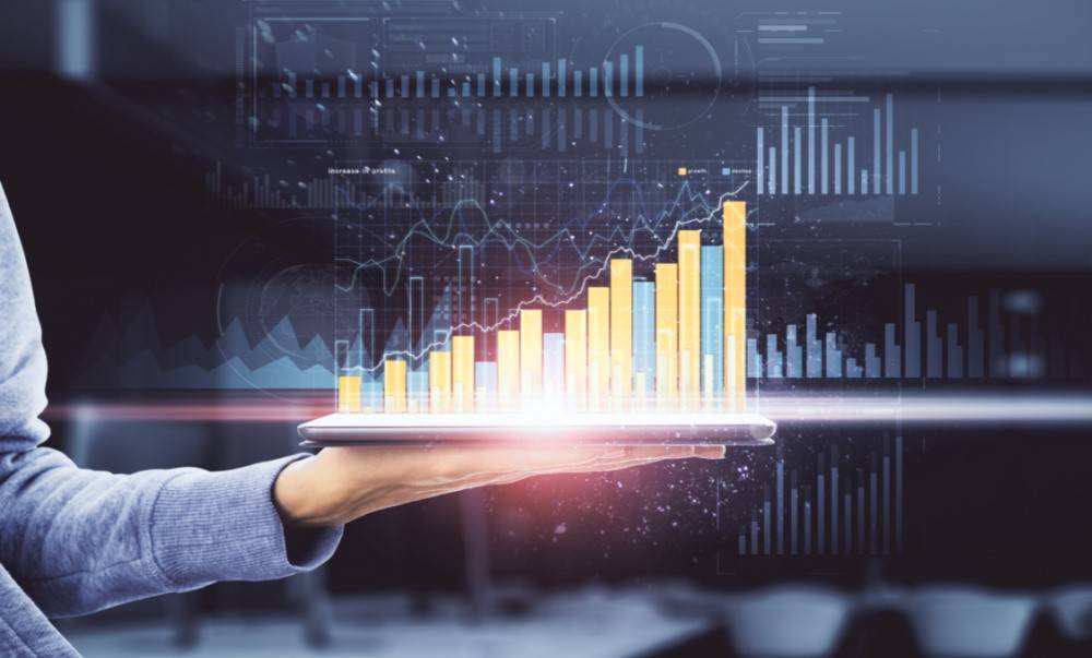 Hand holding a tablet displaying a rising bar chart and digital analytics dashboard, representing business growth, data analysis, and performance tracking.