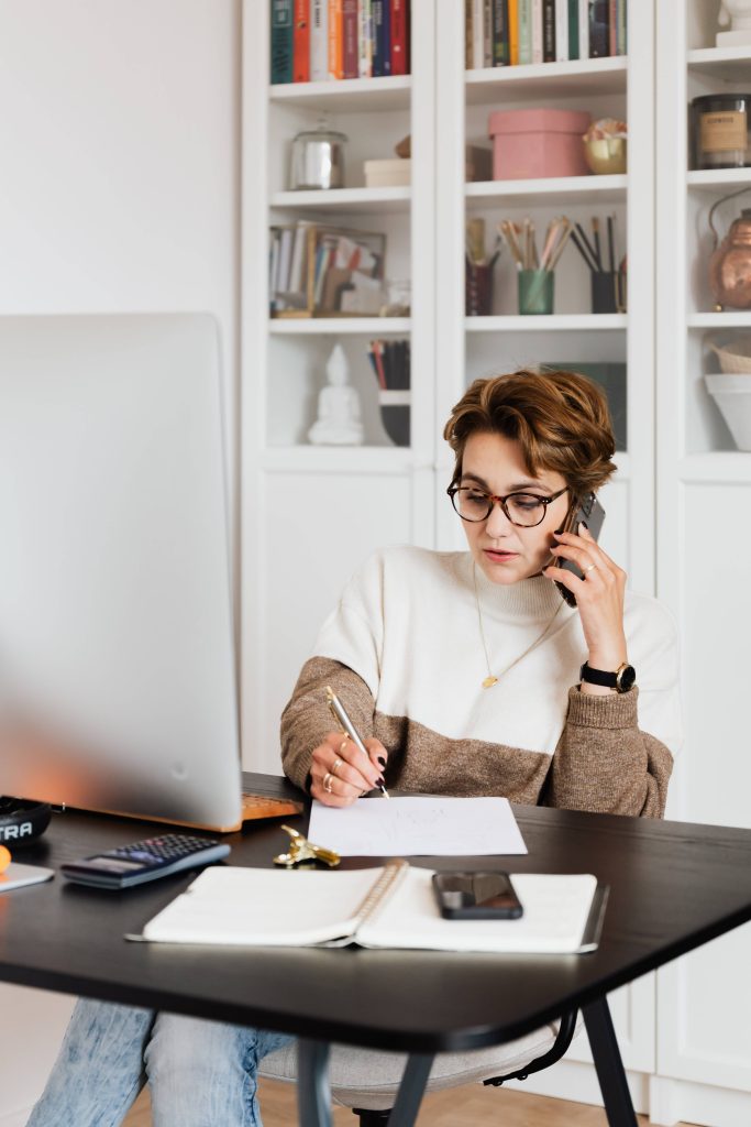 A woman wearing glasses talks on a phone while writing notes at a desk with a laptop, notebook, and smartphone in a bright home office with bookshelves in the background.