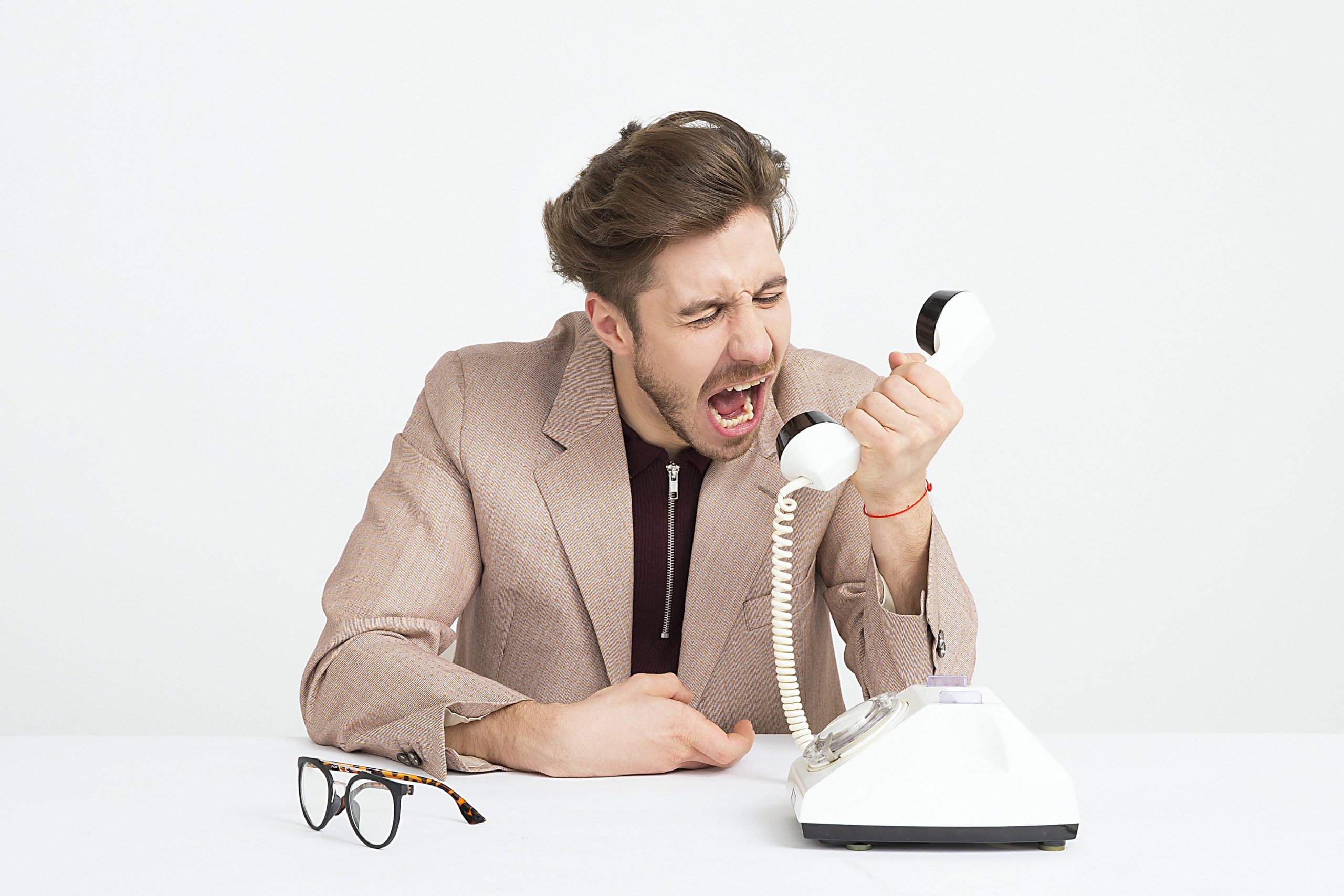 A frustrated man in a blazer shouting into a vintage corded telephone at a desk, expressing anger or stress during a tense phone call.