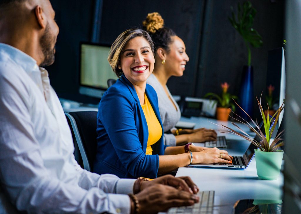 Three coworkers sitting at desks in a modern office, smiling and working on computers, conveying teamwork, collaboration, and a positive professional environment.