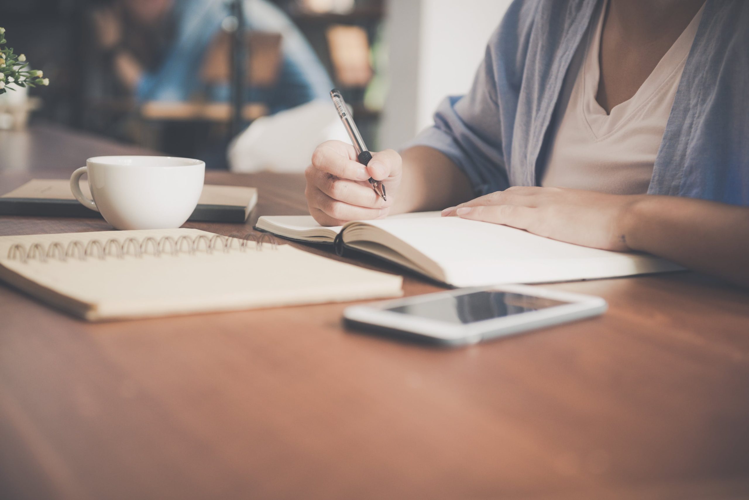 Person writing in a notebook at a desk with a coffee cup, smartphone, and planner nearby, representing focused work, note-taking, and a modern workspace.