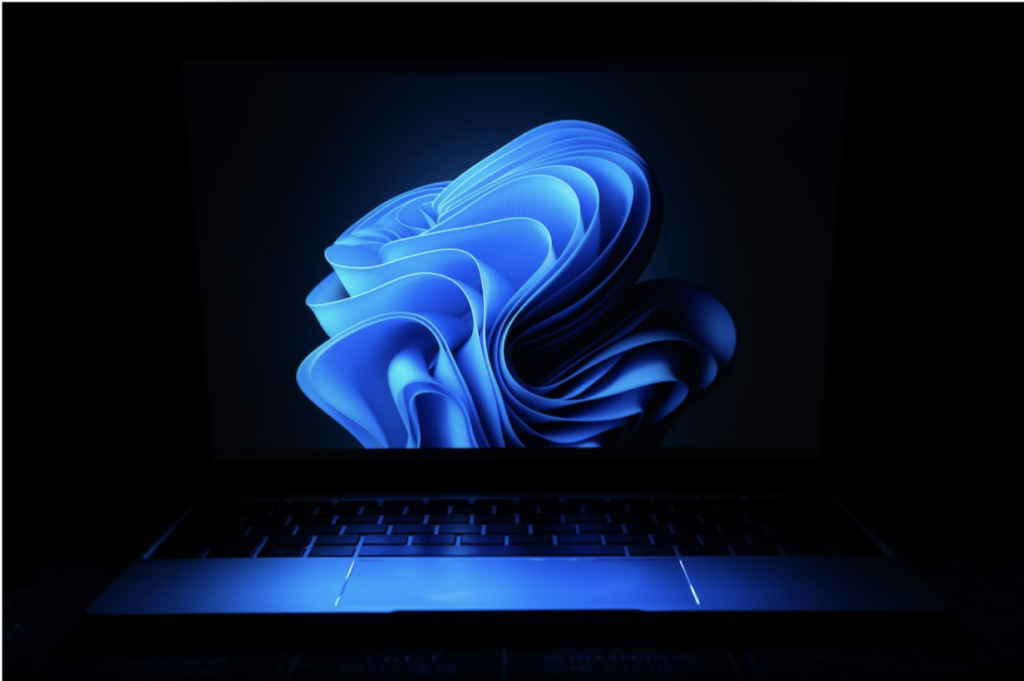 Laptop in a dark setting displaying a glowing blue abstract swirl on the screen, representing a modern operating system interface or advanced digital technology.