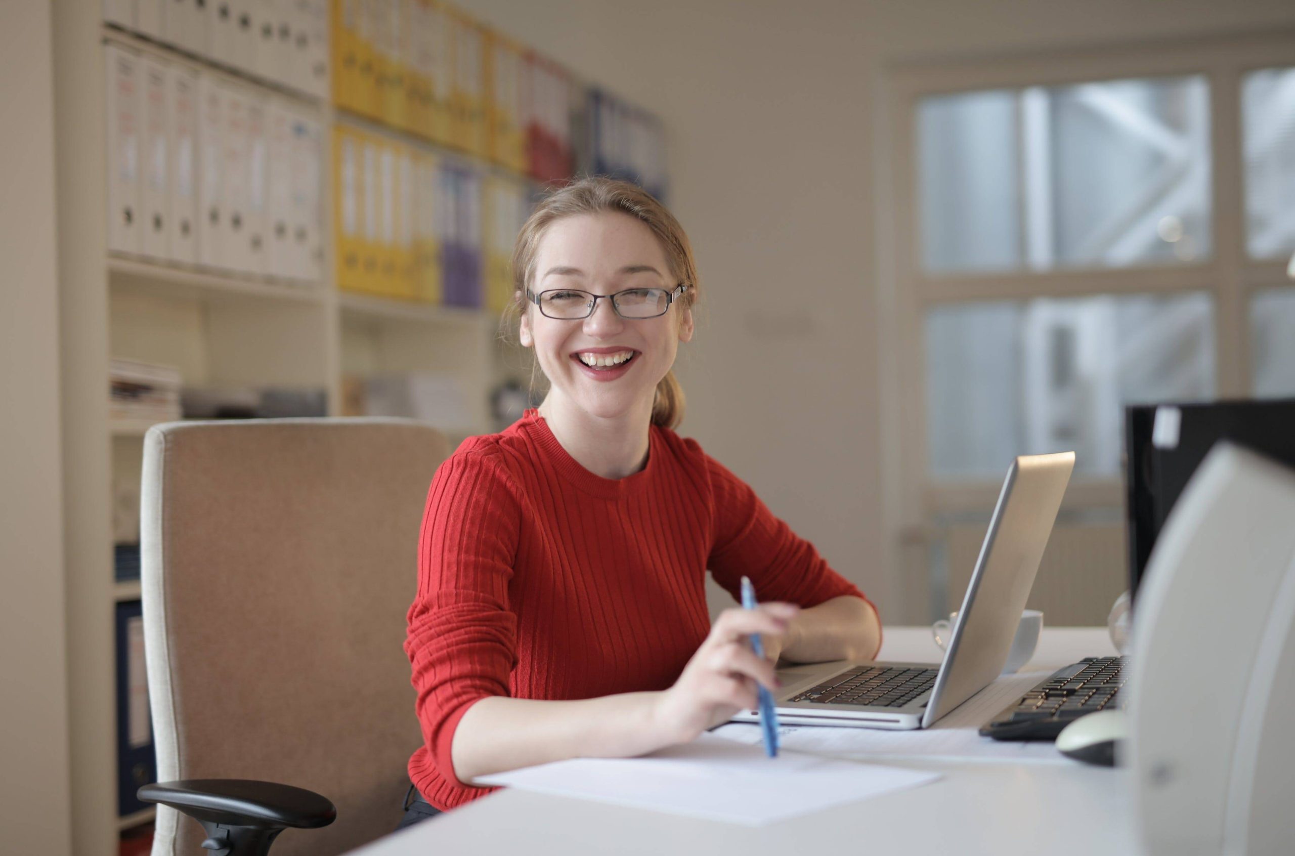 Smiling woman wearing glasses working at a desk with a laptop and pen in a bright office