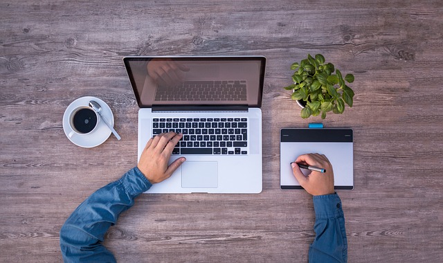 Top-down view of a desk with a laptop, a hand using a drawing tablet, a coffee cup, and a small plant.