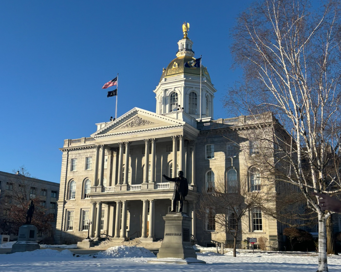 Exterior view of a state capitol building with a gold dome, flags flying, and a statue in front during winter.