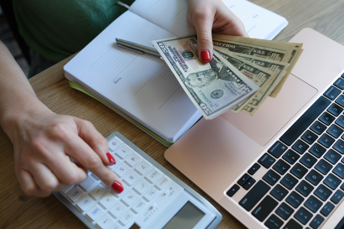 Person counting U.S. dollar bills while using a calculator next to a laptop and notebook.