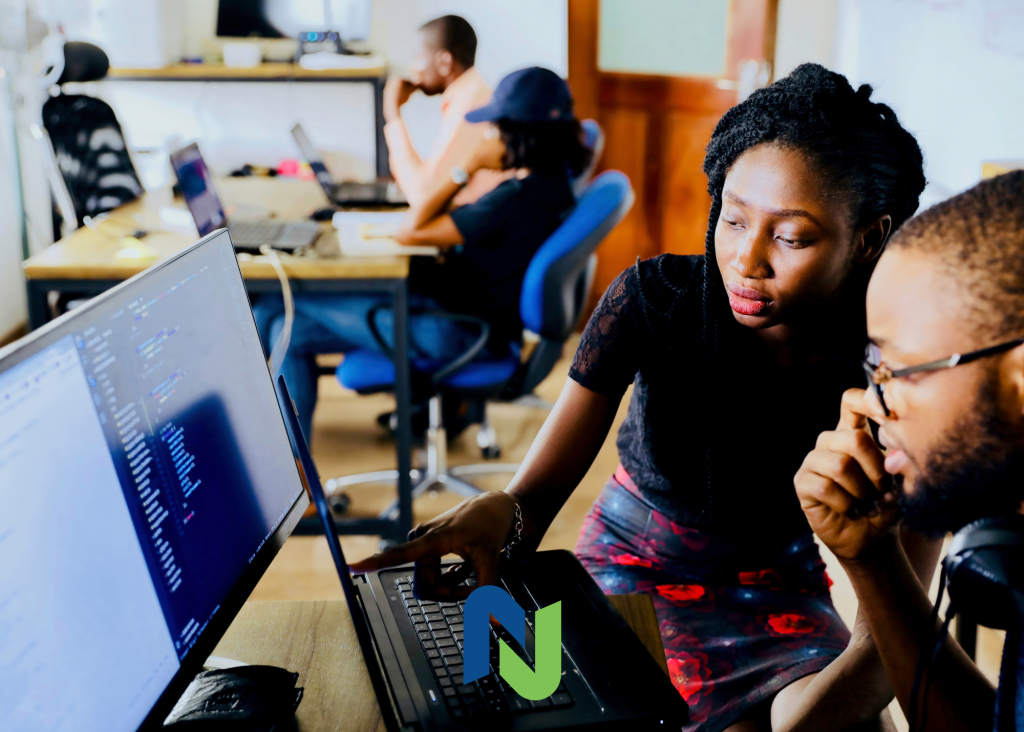 Two coworkers reviewing code on a computer screen in an office while other team members work in the background.